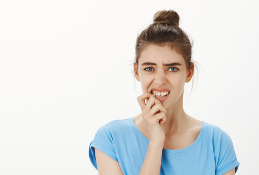 Worried young woman biting her nails, showing uneven teeth and bite concerns, looking anxious on a white background.